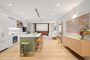 A modern kitchen with a central island, wooden bar stools, a built-in oven, and a dining area with a white sofa and a large abstract painting. - Beachside Apartment in Bondi Wellness Hub (Bondi)