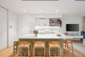 A modern kitchen with a central island, bar stools, and a flat-screen TV. - Beachside Apartment in Bondi Wellness Hub (Bondi)
