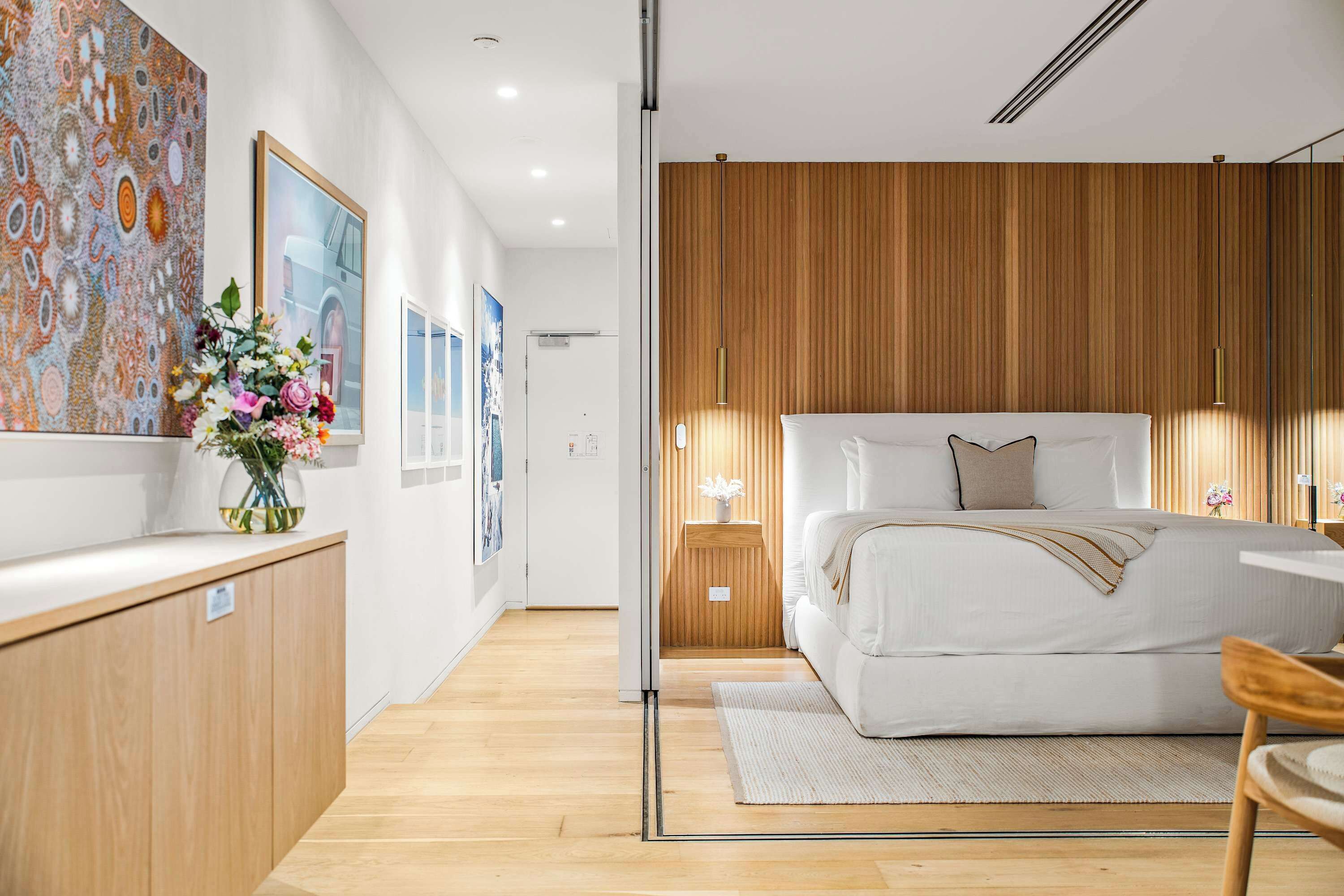 A modern bedroom with a wooden headboard, a bed with white linens, a bedside table, and a wall-mounted light fixture.