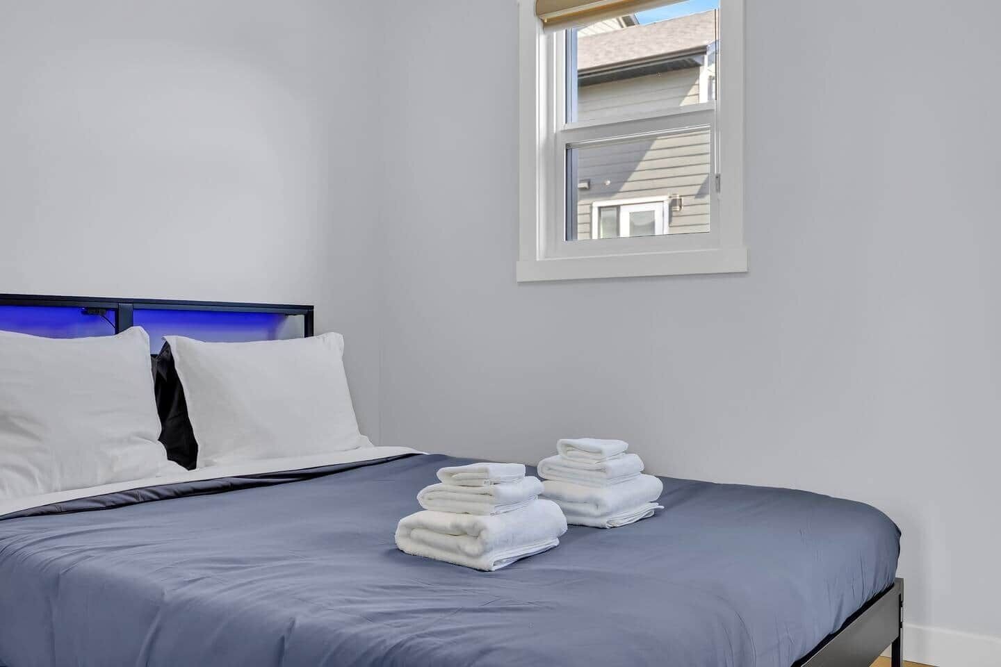 A neatly made bed with white pillows and a blue bedspread, a window with a view of a building, and a stack of folded towels on the bed.