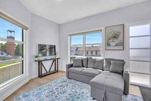 Ein modernes Wohnzimmer mit einem grauen Sofa, einem Fernseher auf einem Holzboden und großen Fenstern mit Blick auf Gebäude und eine Straße.