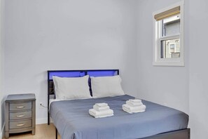 Une chambre à coucher avec un lit, une table de chevet, une fenêtre et une vue sur un bâtiment à l’extérieur.