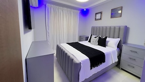 A bedroom with a bed, bedside tables, a TV, a ceiling light, and a window with curtains. - Family Comfort Apt, W/ Patio. BBQ/ Gym (Santiago de los Caballeros)