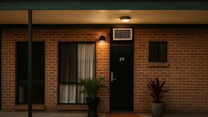 Edificio de ladrillo con puerta negra marcada '24', ventana con cortinas, planta en maceta y unidad de aire acondicionado pequeña sobre la puerta.