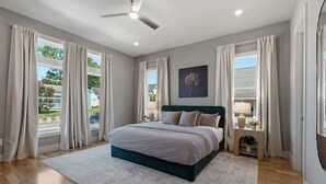 Une chambre à coucher avec un lit, un ventilateur de plafond, une fenêtre avec des rideaux et une table de chevet avec une lampe.