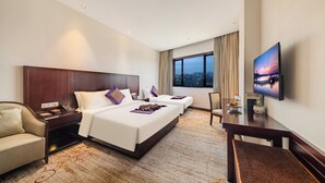 Habitación de hotel con una cama grande, una cama más pequeña, un escritorio, una silla, un televisor y vistas de la ciudad.