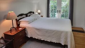 A bedroom with a bed, a nightstand, a lamp, a dresser, a window with curtains, and a view of greenery outside.
