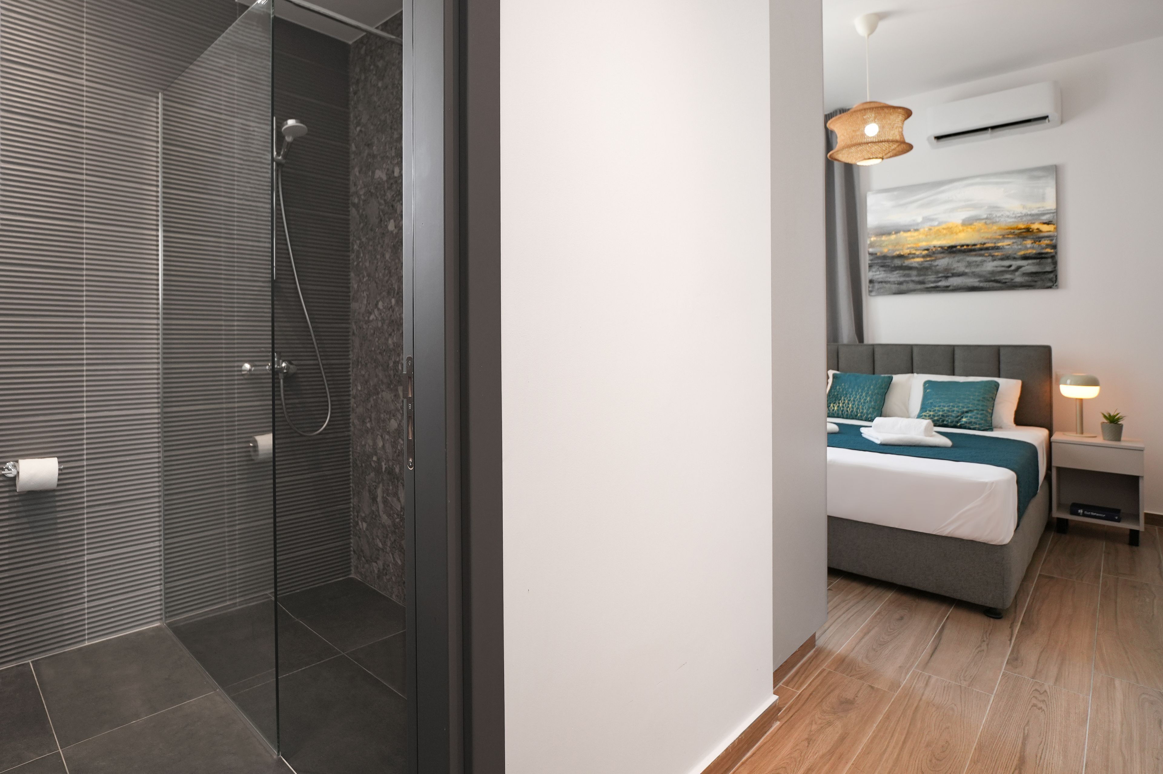 A modern bathroom with a glass shower enclosure and a separate toilet area. The bedroom features a bed with a grey headboard, a bedside table with a lamp, and a wall-mounted air conditioner.