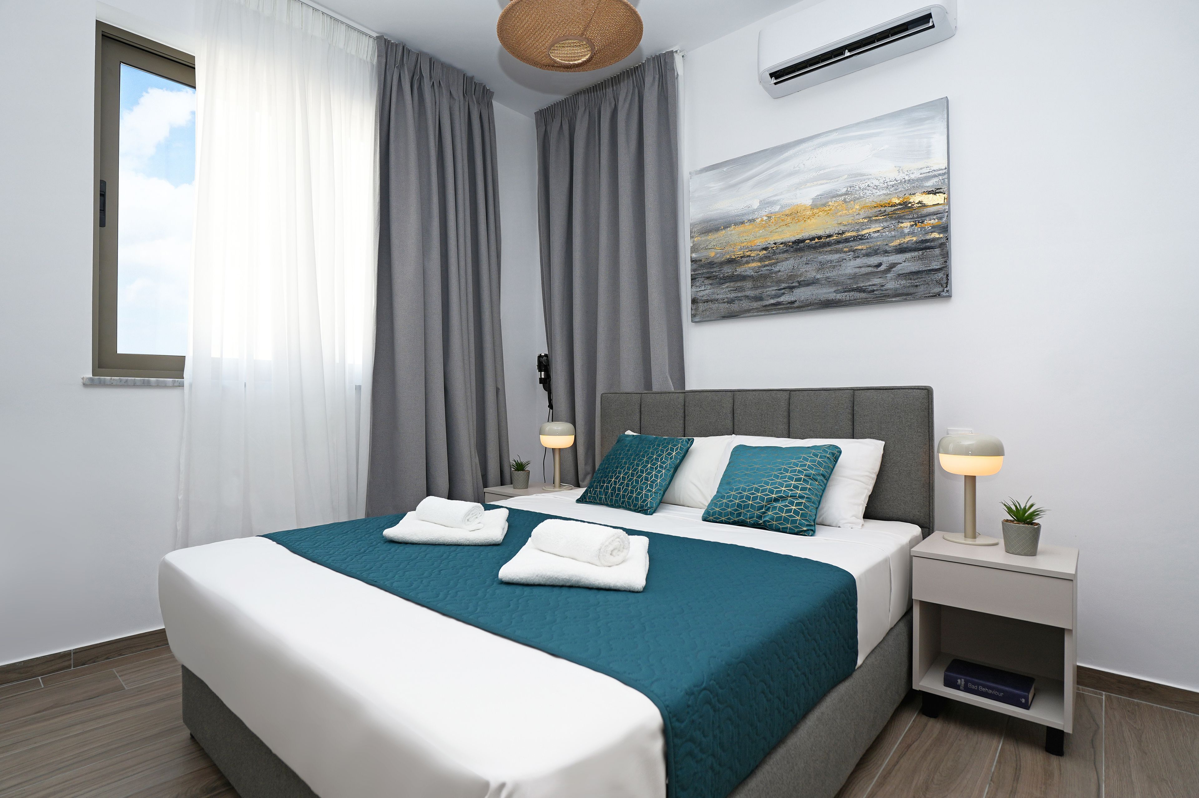 Ein modernes Hotelzimmer mit einem großen Bett, grauen Vorhängen, einer wandmontierten Klimaanlage, einer Nachttischlampe und einem kleinen Topfpflanzen.