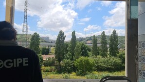 A person in a dark shirt with 'OGIPE' visible, looking out from a window with a view of a hill, trees, and a power line tower. - Lisbon Metro Yellow Line (Loures)