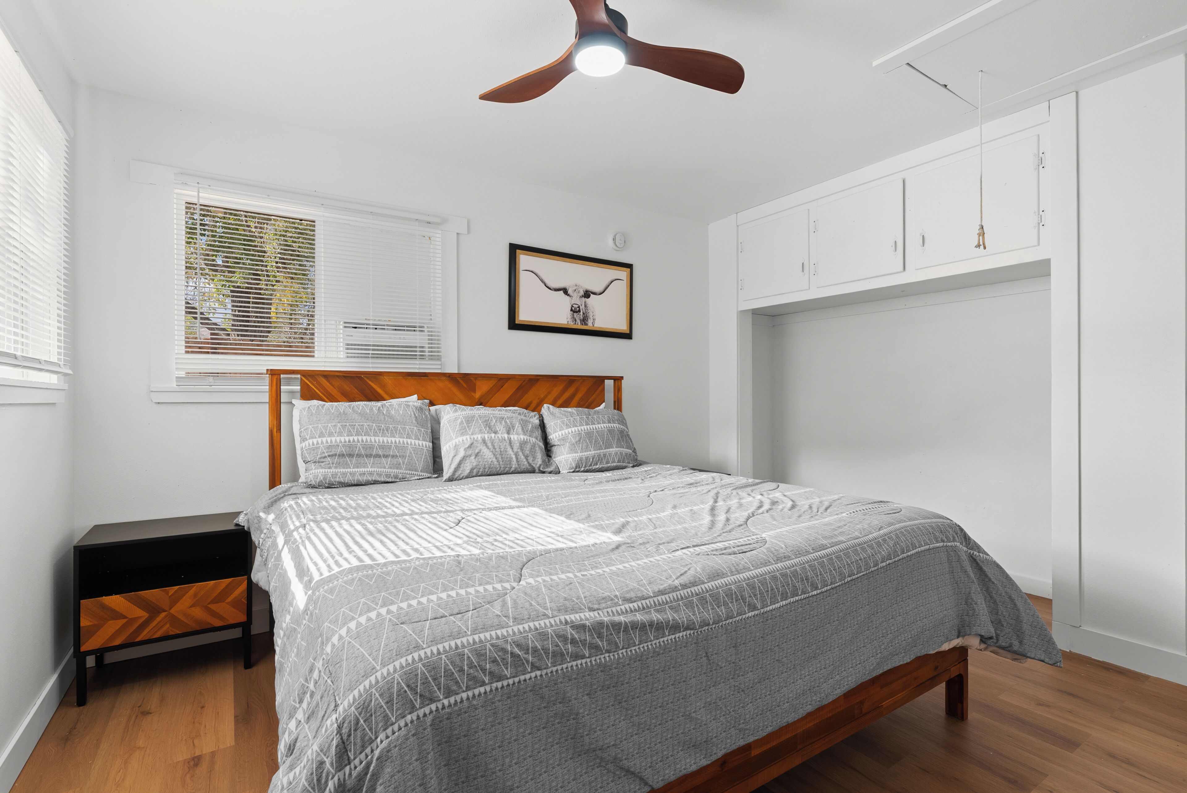 Un dormitorio con cama, mesita de noche, ventilador de techo, ventana con persianas e imagen enmarcada en la pared.