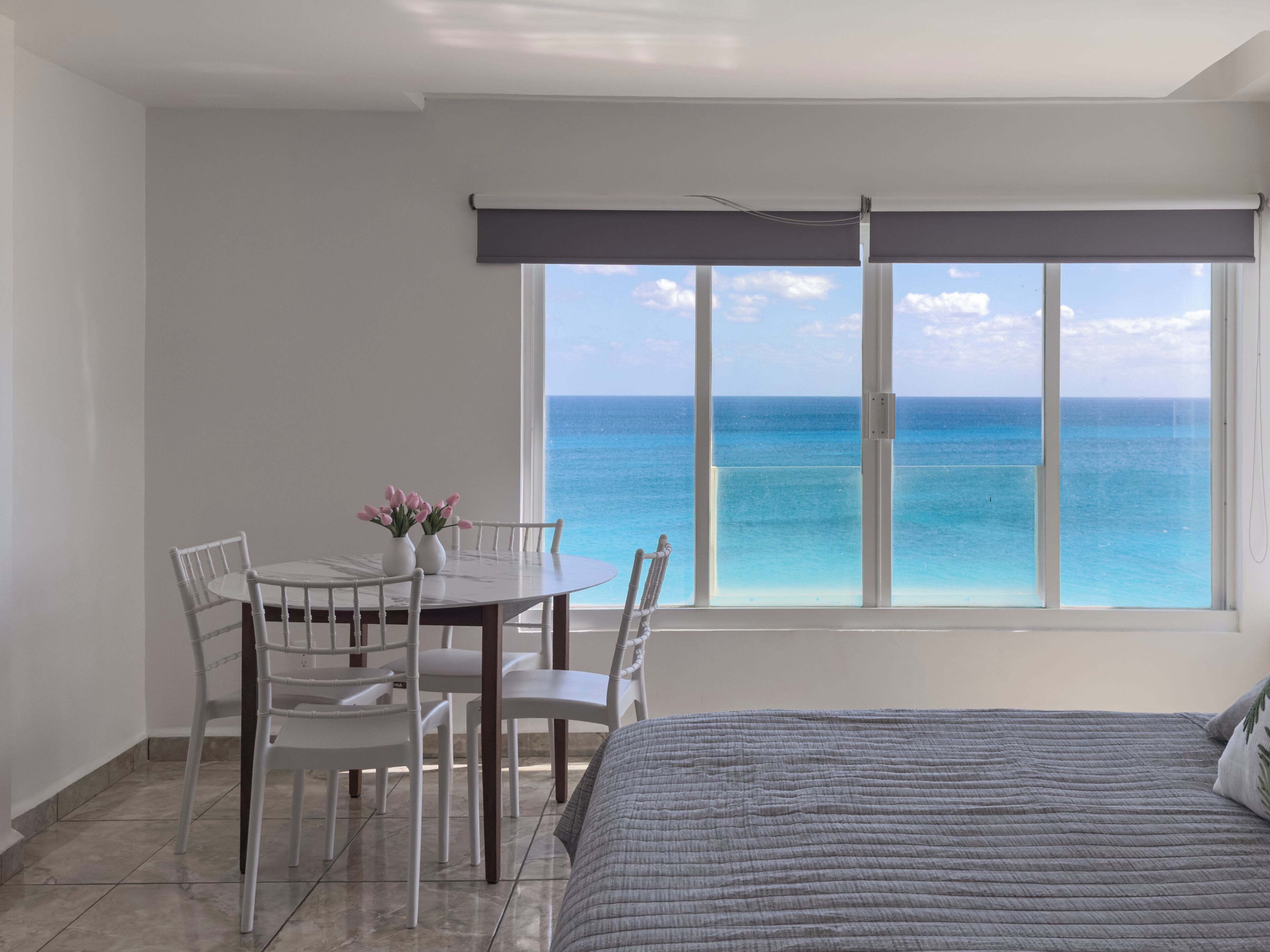A bedroom with a bed, a dining table with chairs, and a view of the ocean through large windows.