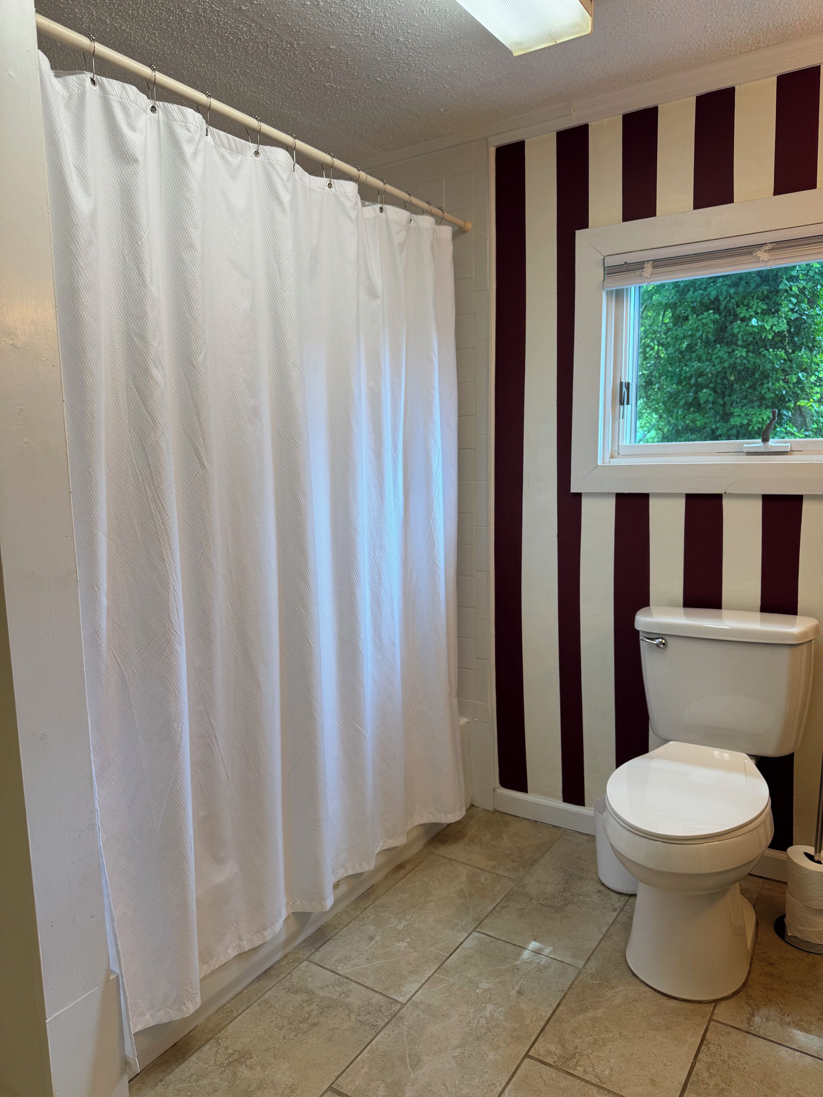 A bathroom with a white shower curtain, a toilet, and a window with a view of trees.