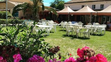 Zona salotto all'aperto con tavoli e sedie bianche, circondata da piante e fiori.