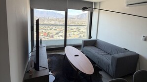 En moderne stue med sofa, sofabord, fjernsyn og et stort vindue med udsigt over byen.