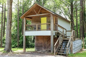 A wooden cabin with a balcony, a yellow door, and a staircase leading to a covered area. - “The Loft on New Natchitoches” (West Monroe)