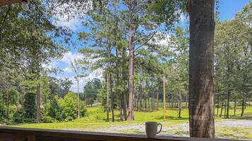 A view from a wooden deck with a white mug on the railing, overlooking a grassy area with trees and a clear sky.