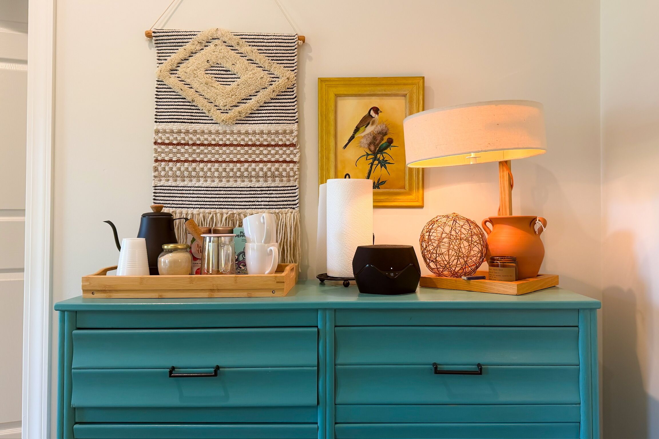 A teal dresser with drawers, a macrame wall hanging, a framed bird painting, a lamp, and various decorative items.