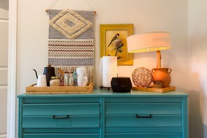 A teal dresser with drawers, a macrame wall hanging, a framed bird painting, a lamp, and various decorative items. - “The Loft on New Natchitoches” (West Monroe)