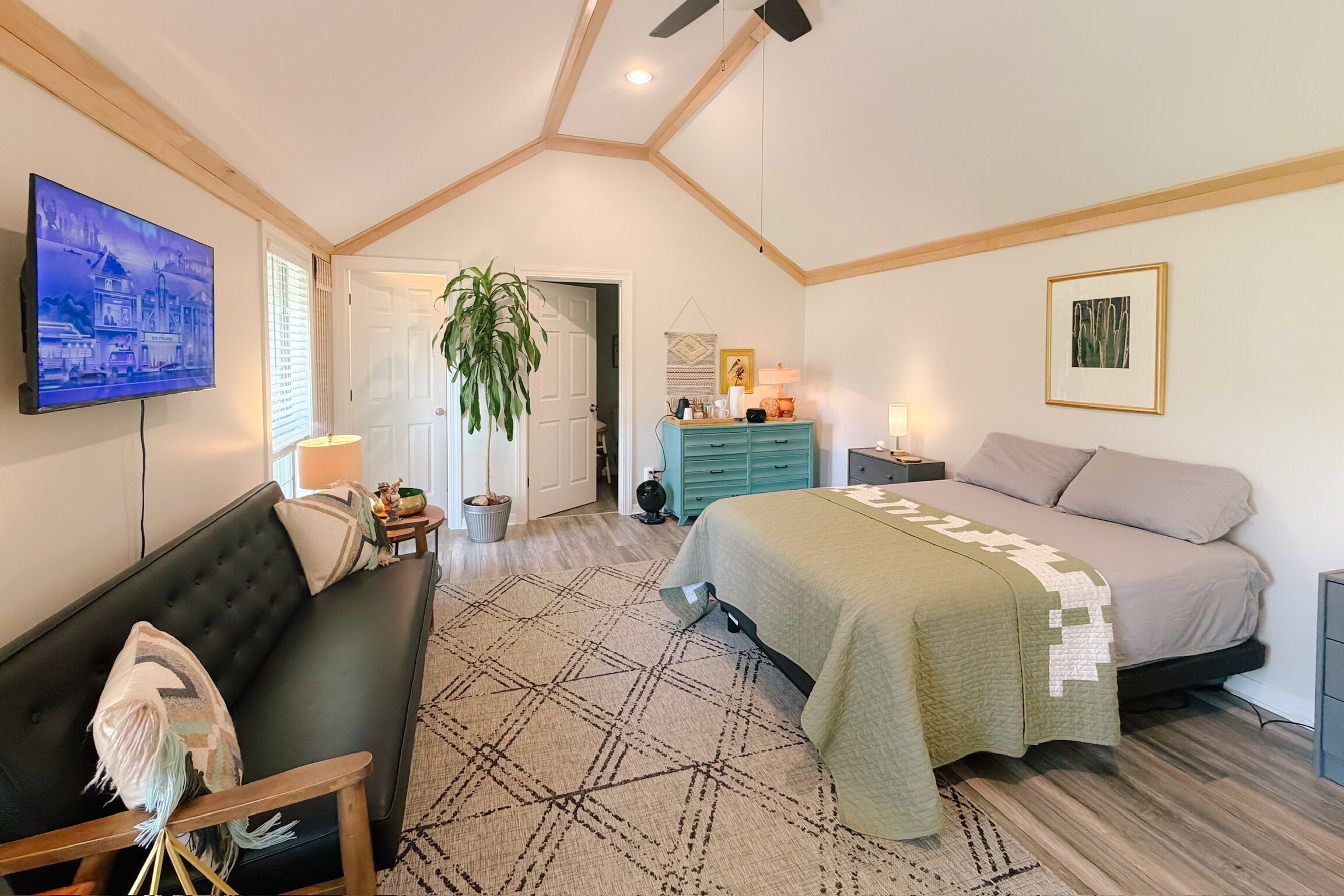 A bedroom with a bed, a sofa, a TV, a ceiling fan, and a door leading to another room.