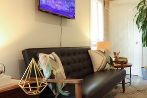 A living room with a dark leather sofa, a mounted television, a small round table with a lamp, and a potted plant. - “The Loft on New Natchitoches” (West Monroe)