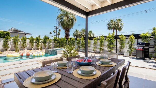 En udendørs terrasse med swimmingpool, spisebord dækket med tallerkener og glas, og en grill.