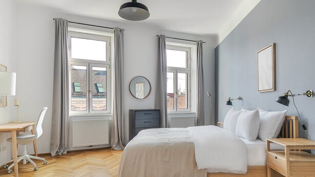 Ett sovrum med en säng, ett skrivbord, en stol och två fönster med gardiner.