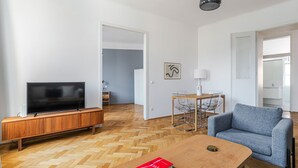 Ett modernt vardagsrum med en platt-TV, ett träskåp, ett parkettgolv i fiskbensmönster, en blå fåtölj, ett soffbord och en matplats med ett bord och stolar.