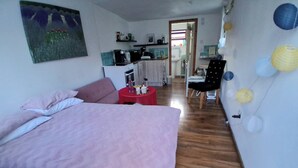 A bedroom with a bed, a small red table, a chair, and a kitchenette in the background.