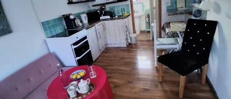 A compact kitchen with a refrigerator, stove, and sink. A dining area with a table set for tea and a black upholstered chair.