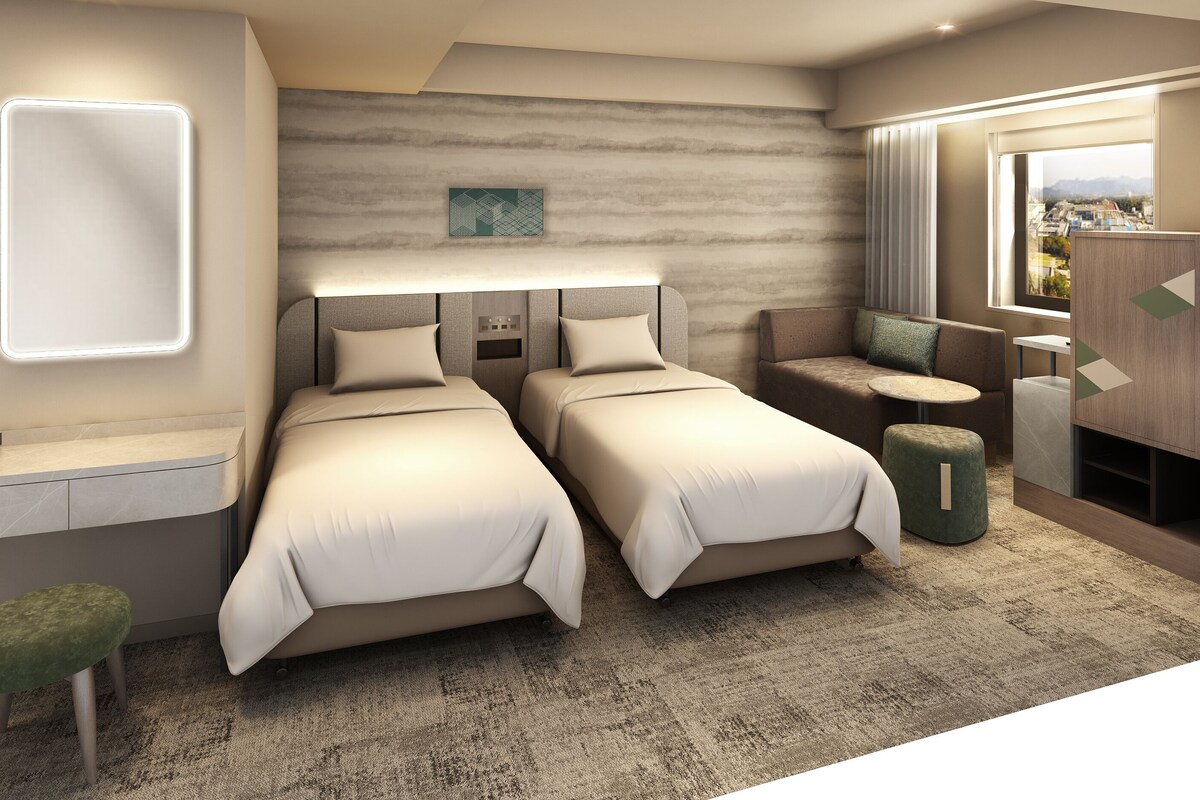 Une chambre d’hôtel moderne avec deux lits, un bureau, un petit coin salon et une vue sur la ville.