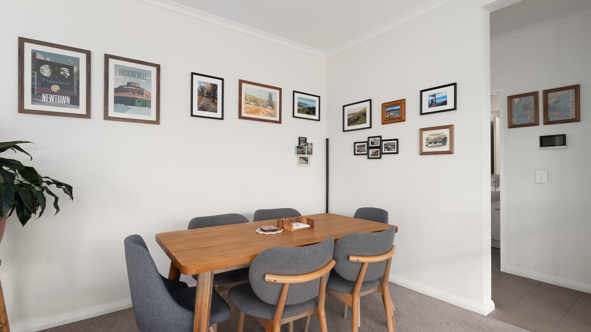 A dining area with a wooden table and grey upholstered chairs, framed pictures on the wall, and a potted plant.