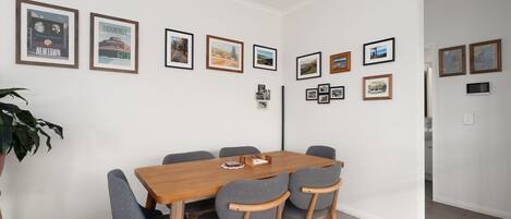 A dining area with a wooden table and grey upholstered chairs, framed pictures on the wall, and a potted plant.