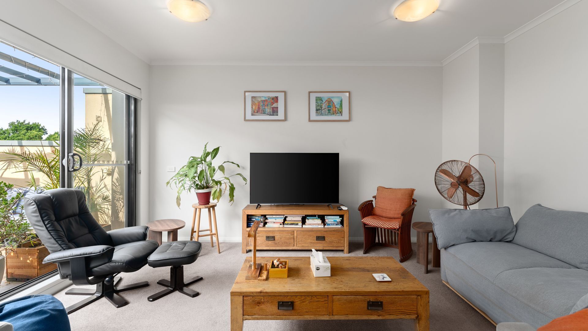A modern living room with a sofa, a black leather chair, a wooden coffee table, a TV stand, and a fan.