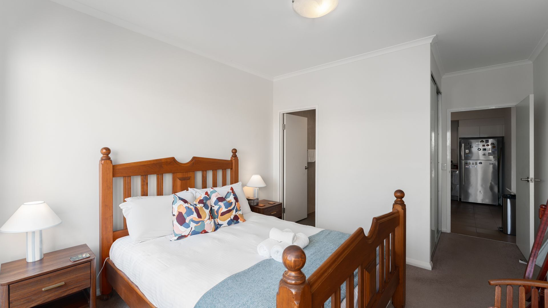 A bedroom with a wooden bed, bedside tables, a lamp, and a view into the kitchen.