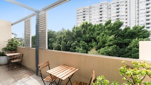 Un balcó amb taula i caires, una planta en test, i vista d'un edifici i arbres.