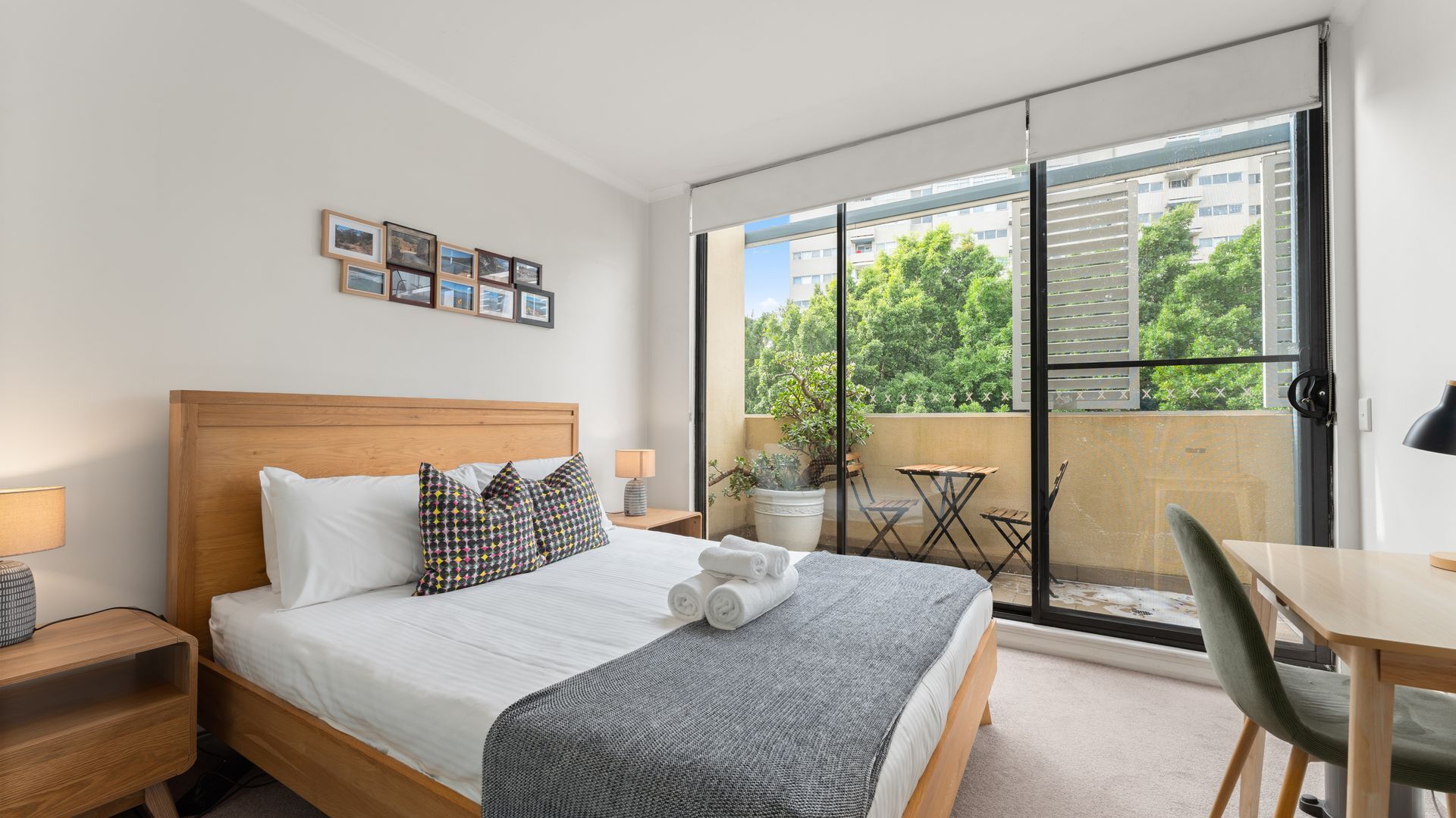 A hotel room with a bed, a desk, a chair, a balcony with a table and chairs, and a potted plant.