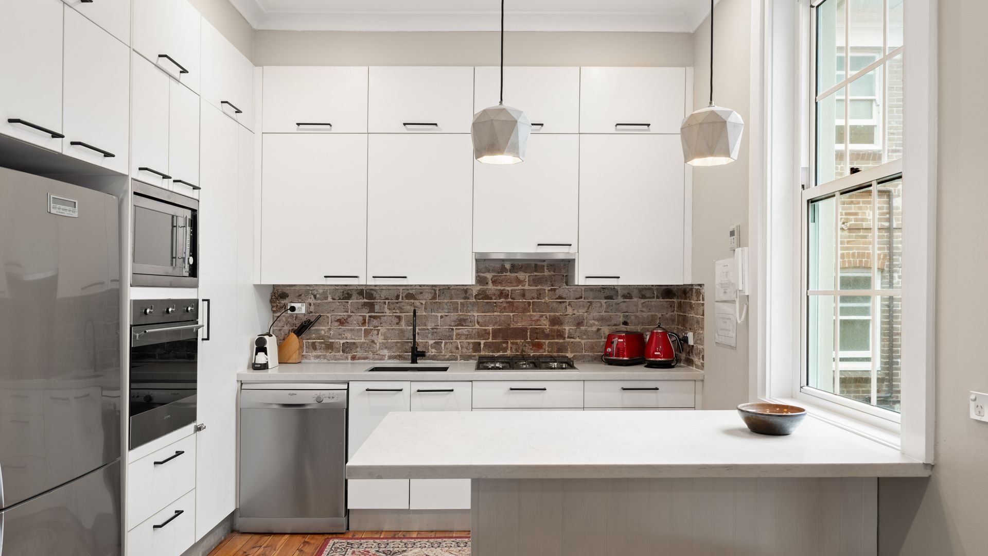 A modern kitchen with white cabinets, a brick backsplash, stainless steel appliances, and pendant lighting. - The Coastal Quarter at Coogee (Coogee)