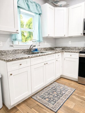 A kitchen with white cabinets, a granite countertop, a window with a view of greenery, and a patterned rug on the floor. - NEW!!! Cozy 2-bedroom house minutes to historic downtown St. Augustine & ocean. (St. Augustine)