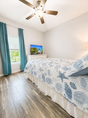 Un dormitorio con piso de madera, una cama con colcha azul y blanca estampada, un ventilador de techo, una ventana con cortinas y un televisor sobre un tocador.