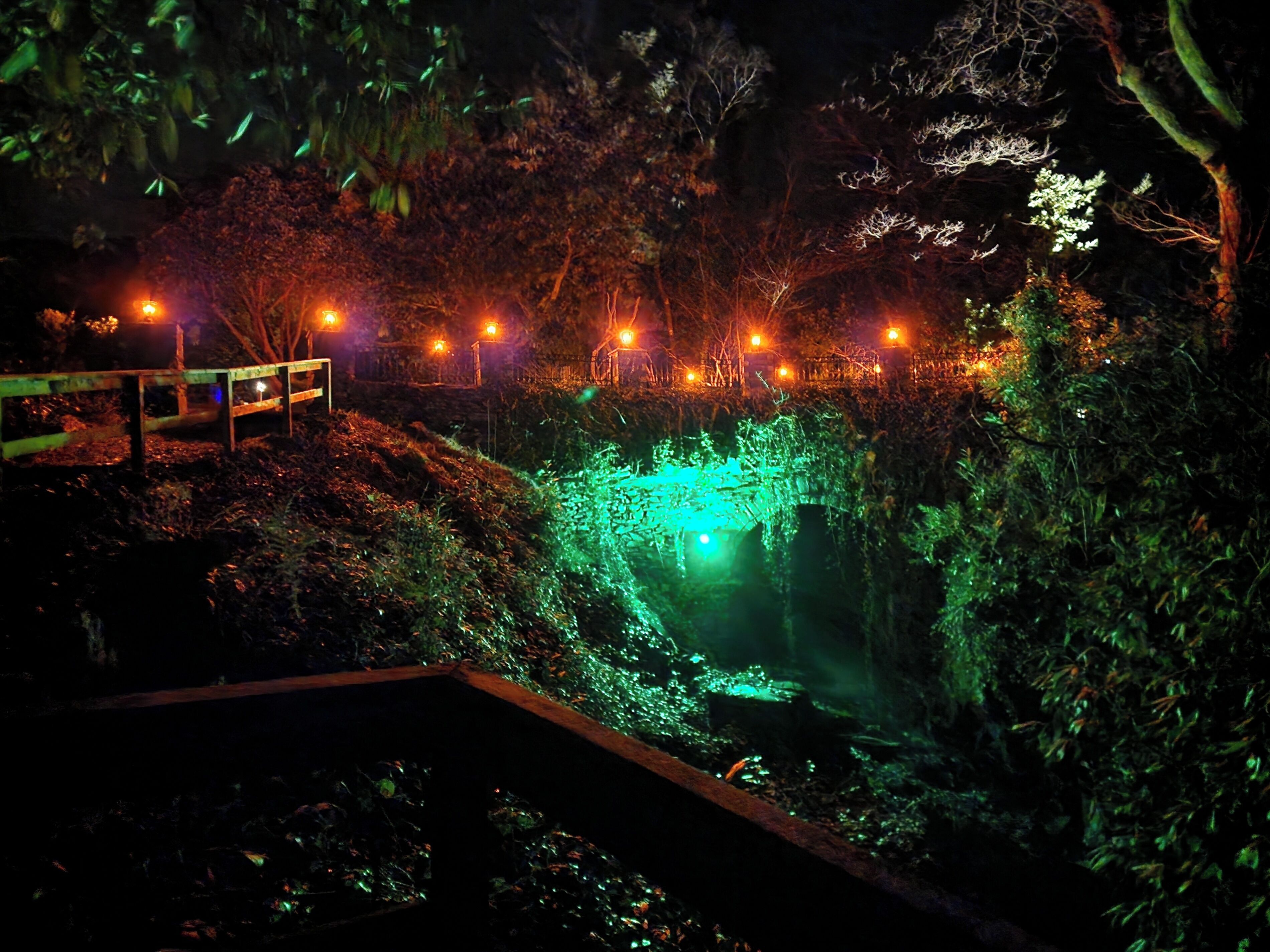 A nighttime view of a park with a bridge, illuminated by warm lights, and a green-lit water feature.