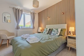 Ein Schlafzimmer mit Bett, Nachttischen, einem Sessel, einem Fenster mit Vorhängen und einer Wand mit Holzlatten.