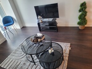 Un soggiorno con televisore, una poltrona blu, un tavolino rotondo in vetro con candele, una piccola pianta e un tappeto decorato.