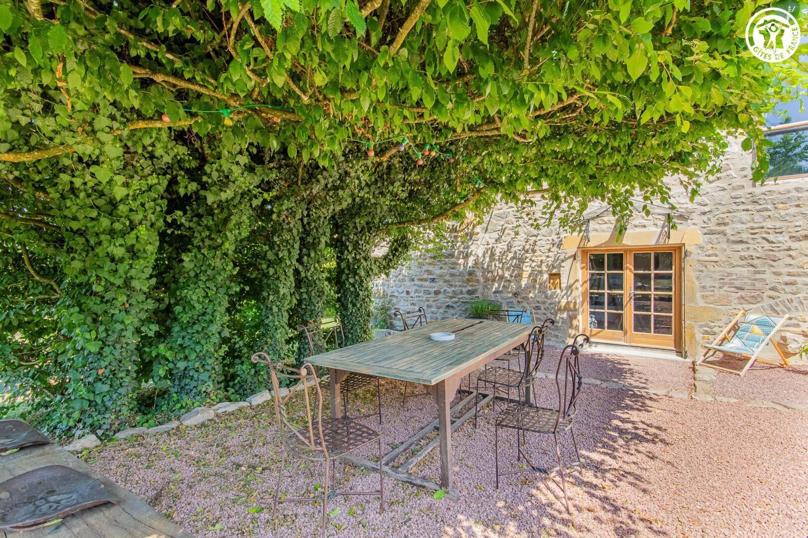 A stone building with a wooden table and chairs under a vine-covered tree.