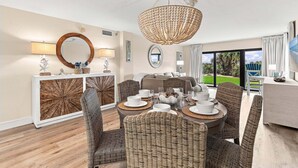 Un comedor con sillas de mimbre, una mesa redonda con platos y tazas blancos, una lámpara de araña grande y vistas al exterior a través de amplios ventanales.