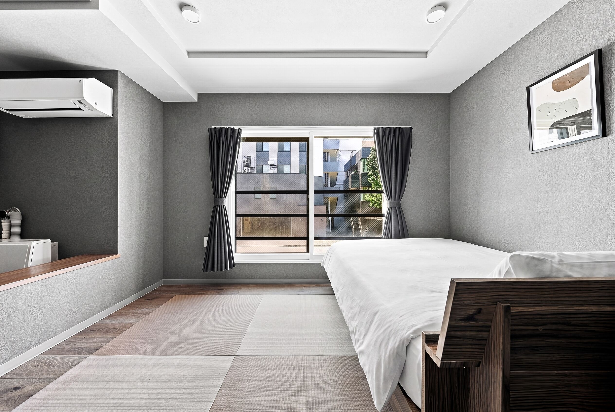 A modern bedroom with a large window, a bed, a bedside table, and a wall-mounted air conditioning unit.
