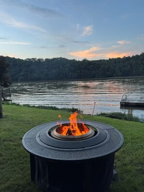 Ein runder Feuerkorb im Freien mit Flammen, umgeben von Gras, mit Blick auf einen stillen See mit Steg und Bäumen im Hintergrund.