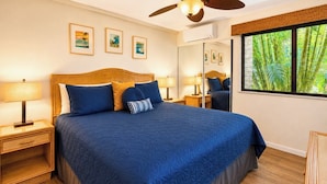 A bedroom with a large bed, wooden headboard, two bedside tables with lamps, a ceiling fan, and a window with a view of greenery. - Napili Point | Oceanfront 1 BR | Car Incl. w/ 6+ Nights | NAP-B28 by KBM (Lahaina)