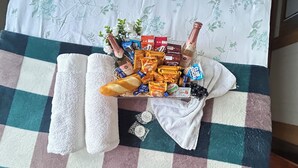 Una cama con colcha a cuadros, una toalla blanca enrollada y una cesta con diversos artículos, incluyendo bombones, una botella de champán y un ramo de flores.