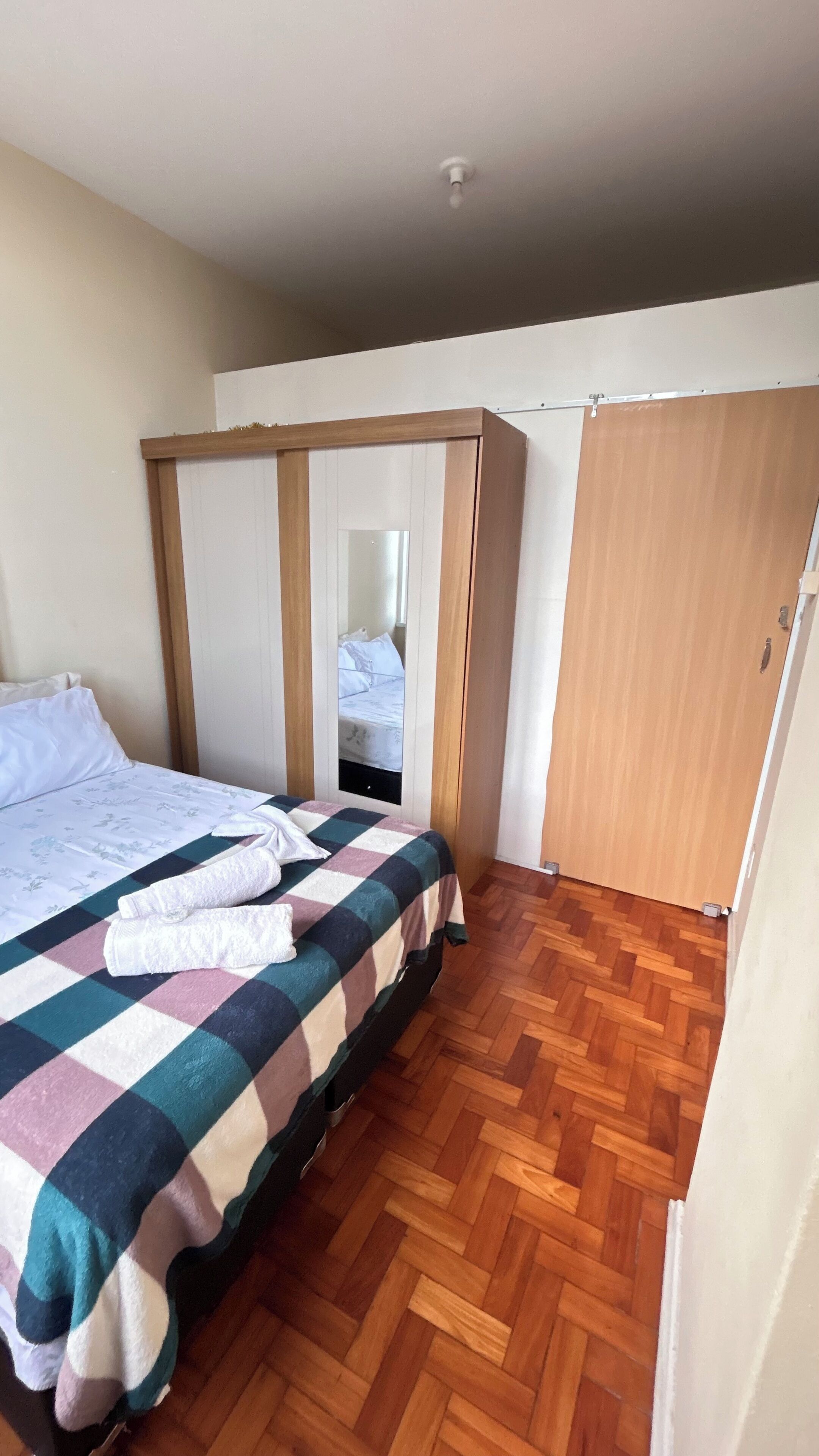 A bedroom with a bed, a wooden wardrobe, a mirror, and a wooden floor.